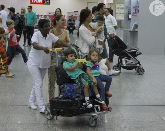 Juliana Paes segura o filho caçula no colo, enquanto o primogênito, Pedro, está sentado no carrinho do aeroporto