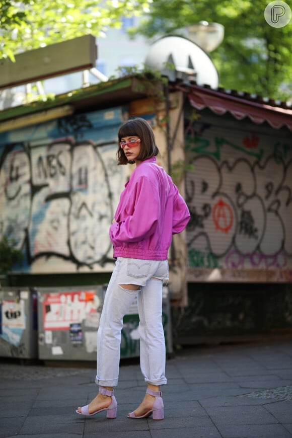 Para um look mais fashion, calça jeans reta clara com recotes abaixo dos bolsos. O toque de cor fica com o casaco pink e as sandálias lilás de salto quadrado