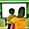 Flavia Sampaio, namorada do empresário Eike Batista, aparece de costas com o filho, Balder, diante da TV e usando a camisa verde e amarela: 'Ainda tinha esperancas. E agora??!!!'