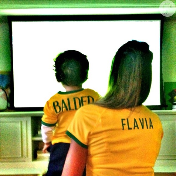 Flavia Sampaio, namorada do empresário Eike Batista, aparece de costas com o filho, Balder, diante da TV e usando a camisa verde e amarela: 'Ainda tinha esperancas. E agora??!!!'