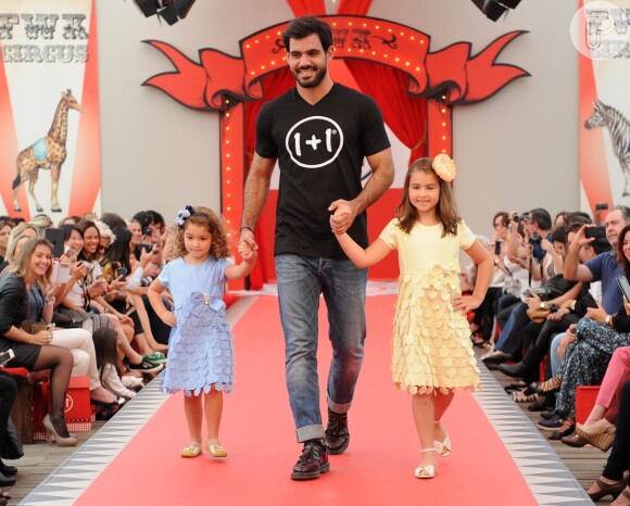 Juliano Cazarré desfila ao lado de duas meninas. O ator vestiu calça jeans e camiseta preta