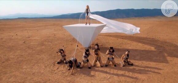 Em outra cena, Britney está em uma plataforma branca seguranda sua bailarinas por correntes