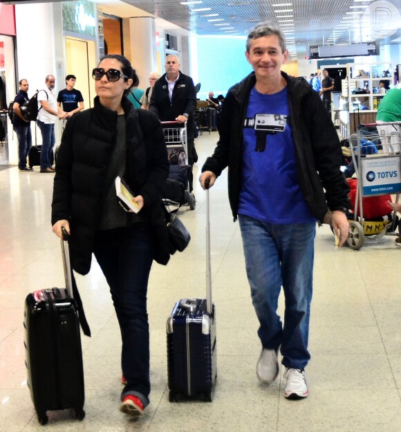 Serginho Groisman caminha sorridente pelo aeroporto com a mulher, Fernanda Molina