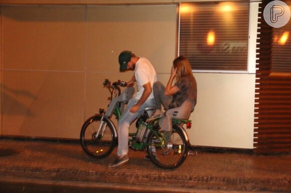 Fernanda Lima, com a mão no rosto, é carregada na garupa da bicicleta do marido, Rodrigo Hilbert