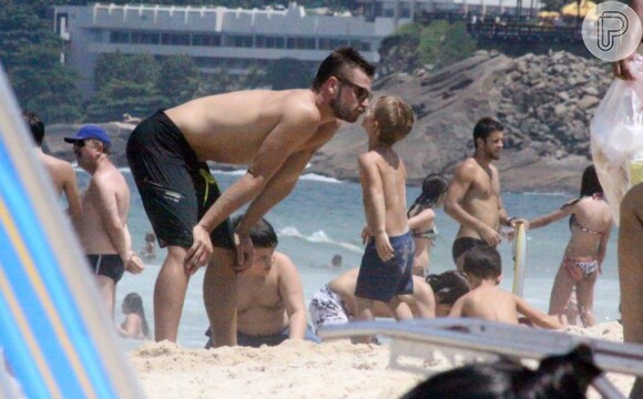 Rodrigo Hilbert se abaixa para ganhar um beijo do filho, na praia do Leblon