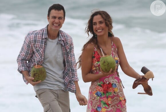 Camila Pitanga e Gabriel Braga Nunes sorriem ao se aproximarem do mar, evitando molhar as roupas do figurino