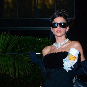 Bruna Marquezine surge em look composto por vestido preto míni com decote tomara que caia, meia calça, luvas, salto alto, óculos escuros e bolsa na mesma cor