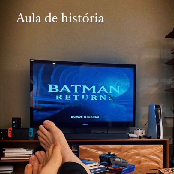 Lucas Lima aparece com os pés encostados aos pés de uma pessoa que não foi identificada. Os dois estão diante de uma televisão que exibe o filme 'Batman: O Retorno'