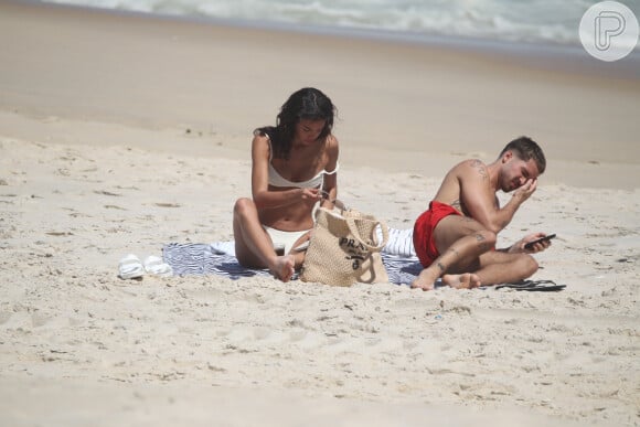 Bruna Marquezine e João Guilherme se deitaram na areia. Ela escolheu ler um livro, enquanto ele mexia no celular