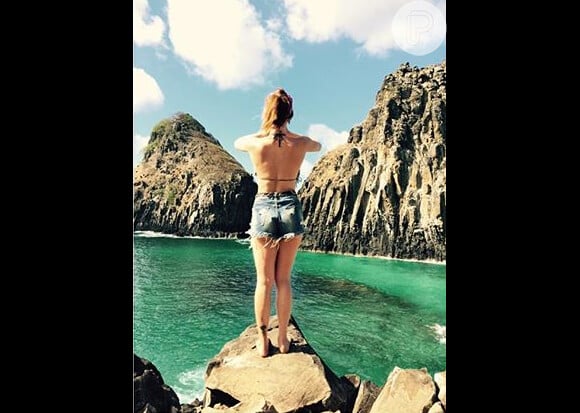 A atriz postou uma foto em cima de uma pedra com a praia de água cristalinas ao fundo