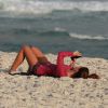 Mariana Goldfarb mexe no celular, deitada na areia, enquanto Cauã Reymond surfa