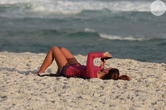 Mariana Goldfarb mexe no celular, deitada na areia, enquanto Cauã Reymond surfa