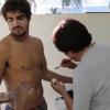Caio Castro observa a maquiadora que vai passar óleo no corpo do ator