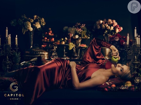 A atriz posa sensualmente em uma mesa de frutas com um vestido vermelho de um ombro só