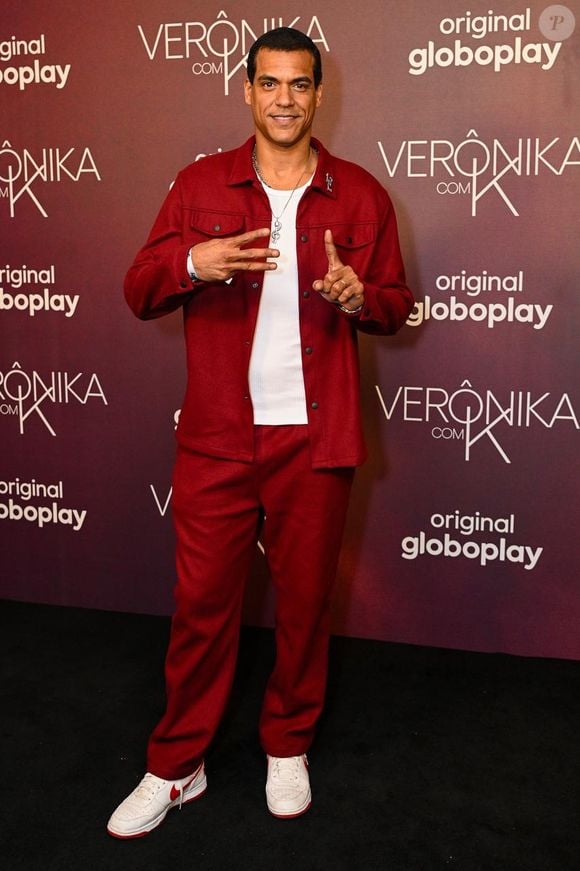 Marcello Melo Jr. vestiu um conjunto de alfaiataria casual em tom vermelho-fechado/bordô, composto por jaqueta estilo overshirt com bolsos no peito e calça de corte reto. Por baixo da jaqueta aberta, utiliza uma camiseta regata canelada branca. O visual urbano e esportivo é completado por tênis brancos com o logotipo da marca em tom vermelho
