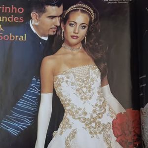 10. Vestido Tomara que caia

Aqui, temos uma noiva mais sensual e glamourosa, mas sem perder a grandiosidade da época. O vestido é tomara que caia com bordados dourados em arabescos florais que descem pelo corpete e se espalham pela saia em forma de cascata, criando um efeito de movimento e leveza.

A saia rodada carrega a tradição volumosa, mas o desenho do bordado alonga e valoriza a silhueta. As luvas longas brancas adicionam um toque clássico, enquanto o styling traz um colar e tiara dourados, em um mood mais minimalista em comparação aos outros dois. O buquê vermelho contrasta de forma ousada com o dourado e o branco, trazendo um impacto visual típico das noivas ousadas do início dos anos 2000.
