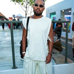 O ator Mussunzinho apostou em uma estética contemporânea de volume arquitetônico, vestindo um conjunto monocromático em tom off-white. O visual é composto por uma túnica sem mangas de corte reto e amarrações laterais, sobreposta a uma calça de modelagem extra-wide com pregas frontais profundas que conferem movimento e fluidez