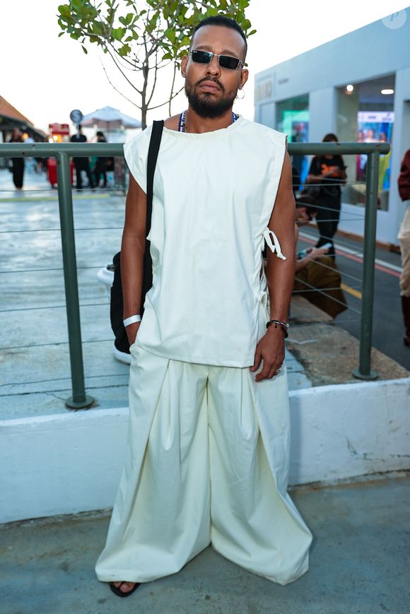 O ator Mussunzinho apostou em uma estética contemporânea de volume arquitetônico, vestindo um conjunto monocromático em tom off-white. O visual é composto por uma túnica sem mangas de corte reto e amarrações laterais, sobreposta a uma calça de modelagem extra-wide com pregas frontais profundas que conferem movimento e fluidez