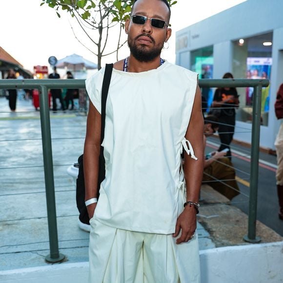 O ator Mussunzinho apostou em uma estética contemporânea de volume arquitetônico, vestindo um conjunto monocromático em tom off-white. O visual é composto por uma túnica sem mangas de corte reto e amarrações laterais, sobreposta a uma calça de modelagem extra-wide com pregas frontais profundas que conferem movimento e fluidez