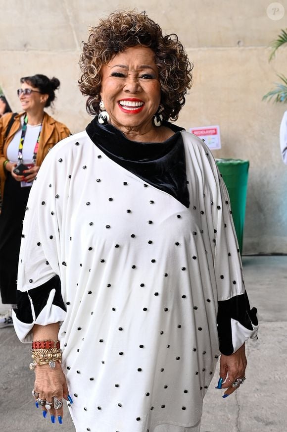 A peça de Alcione no Rio2C, traz uma gola assimétrica imponente em veludo molhado preto, textura que se repete nos punhos dramáticos e equilibra o volume da túnica, com uma calça pantalona de caimento impecável e sapatos de bico fino pretos com aplicações de tachas