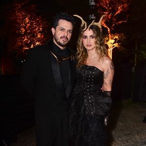 Mari Palma e marido em looks coordenados em preto. Ela usa um vestido de festa preto tomara que caia com corpete estruturado por barbatanas e detalhes de ilhós centrais. A saia tem camadas de babados texturizados e transparentes. Ele veste um terno preto clássico com lapela de cetim, sobre uma camisa preta e um detalhe metálico dourado na gola