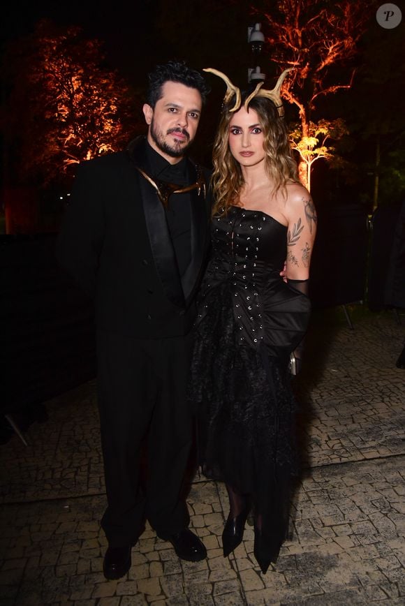 Mari Palma e marido em looks coordenados em preto. Ela usa um vestido de festa preto tomara que caia com corpete estruturado por barbatanas e detalhes de ilhós centrais. A saia tem camadas de babados texturizados e transparentes. Ele veste um terno preto clássico com lapela de cetim, sobre uma camisa preta e um detalhe metálico dourado na gola
