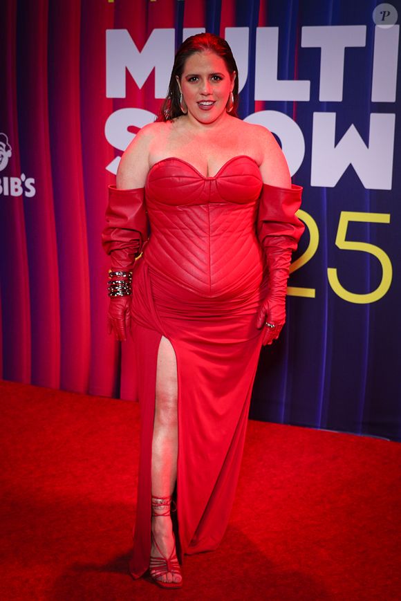 Tati Machado apareceu poderosa em um vestido vermelho intenso, de modelagem estruturada e inspiração quase armadura fashion. A peça une corset acolchoado, mangas longas caídas e uma fenda lateral que traz sensualidade na medida certa. As luvas no mesmo tom e as sandálias de tiras reforçam a estética dramática, enquanto os acessórios metalizados dão um brilho extra