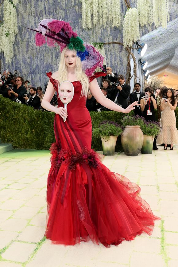 Gwendoline Christie impactou com um vestido sereia vermelho vibrante, rico em camadas de tule e penas na barra, acompanhado por um imenso adereço de cabeça com plumas coloridas e uma máscara de mão clássica