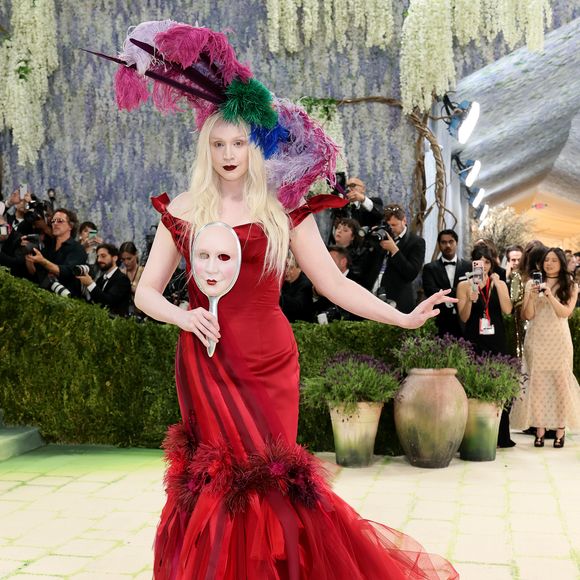 Gwendoline Christie impactou com um vestido sereia vermelho vibrante, rico em camadas de tule e penas na barra, acompanhado por um imenso adereço de cabeça com plumas coloridas e uma máscara de mão clássica