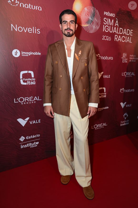 Enzo Celulari exibiu uma produção sofisticada em tons terrosos, composta por um blazer de abotoamento duplo (double-breasted) em marrom chocolate com botões dourados. Ele combinou a peça estruturada com uma calça de alfaiataria em tom creme de caimento impecável e uma regata branca básica por baixo