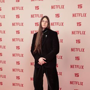 Felca: veste um blazer preto sobre blusa de gola alta escura e calça ampla com brilho aveludado, combinando o conjunto de tom sóbrio com sapato social preto de sola tratorada, corrente no pescoço e óculos de grau finos