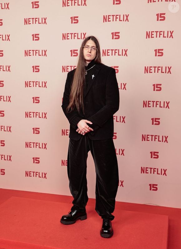 Felca: veste um blazer preto sobre blusa de gola alta escura e calça ampla com brilho aveludado, combinando o conjunto de tom sóbrio com sapato social preto de sola tratorada, corrente no pescoço e óculos de grau finos