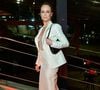 Mariana Ximenes escolheu um terno feminino na cor off-white de acabamento acetinado, composto por blazer de corte alfaiataria acinturado com lapelas pontudas e saia longa ajustada até o chão no mesmo tom. Por baixo do blazer, usa uma peça com transparência e aplicações sutis de brilho. Completa a produção com uma bolsa preta de ombro e cabelos presos em um coque elegante