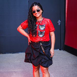 'Tia' Milena: a ex-BBB aposta na estética maximalista com uma t-shirt vermelha inteiramente coberta por paetês e aplicação frontal de estampas de gatos. O visual é complementado por uma saia preta estruturada em babados volumosos, cinto duplo de couro com ilhoses metálicos, óculos retangulares e sapato plataforma preto com meias aparentes