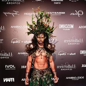 O influenciador Tet veste um traje guerreiro de inspiração botânica sem camisa, composto por uma saia longa feita inteiramente de folhas verdes e aplicações de folhagens e cristais na cintura e no peitoral. O look é coroado por um toucado gigante de plantas e ramagens naturais sobre o cabelo, ombreiras com rostos esculpidos em tom dourado/bronze e braceletes metálicos nos pulsos