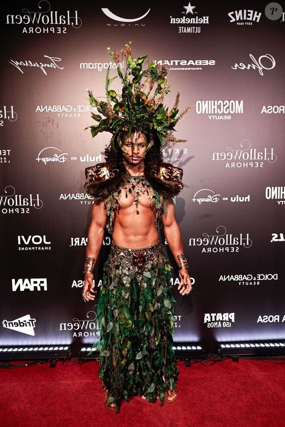 O influenciador Tet veste um traje guerreiro de inspiração botânica sem camisa, composto por uma saia longa feita inteiramente de folhas verdes e aplicações de folhagens e cristais na cintura e no peitoral. O look é coroado por um toucado gigante de plantas e ramagens naturais sobre o cabelo, ombreiras com rostos esculpidos em tom dourado/bronze e braceletes metálicos nos pulsos