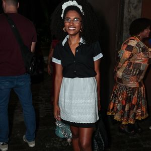 Um traje clássico e delicado de arrumadeira! Mariana Sena usa uma camisa preta de botões com gola e punhos rendados em branco, combinada com uma saia preta e avental branco trabalhado em bordados. No cabelo afro, uma tiara de renda branca com babados dá o toque final ao lado de óculos de grau e tênis estampados