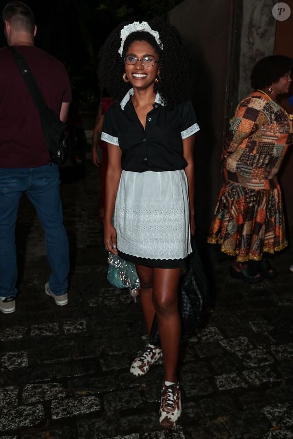 Um traje clássico e delicado de arrumadeira! Mariana Sena usa uma camisa preta de botões com gola e punhos rendados em branco, combinada com uma saia preta e avental branco trabalhado em bordados. No cabelo afro, uma tiara de renda branca com babados dá o toque final ao lado de óculos de grau e tênis estampados