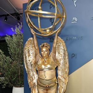 Megan Stalter: apostando em uma caracterização performática e literal da própria estatueta do Emmy, o look monocromático em tom dourado reluzente traz top cropped e hot pants metálicos complementados por imponentes asas de penas esculpidas nas costas. Toda a pele e o cabelo aparecem cobertos por tinta corporal metálica no mesmo tom, emoldurados por colares e cinto de correntes