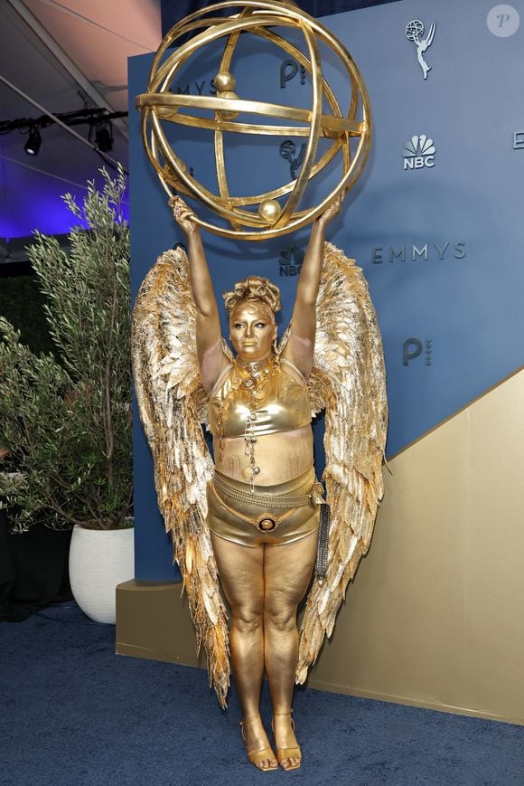 Megan Stalter: apostando em uma caracterização performática e literal da própria estatueta do Emmy, o look monocromático em tom dourado reluzente traz top cropped e hot pants metálicos complementados por imponentes asas de penas esculpidas nas costas. Toda a pele e o cabelo aparecem cobertos por tinta corporal metálica no mesmo tom, emoldurados por colares e cinto de correntes