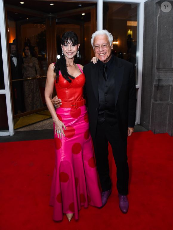 Alexandra Martins e Antônio Fagundes: Alexandra Martins veste um conjunto vibrante e estruturado, composto por um corset de couro vermelho com pespontos aparentes e uma saia longa em tom de rosa choque, decorada com grandes bolas (poás) em vermelho. O look é acentuado por um cinto largo que marca a cintura e brincos de cascata prateados. Ao seu lado, Antônio Fagundes opta por um visual monocromático preto, composto por terno, colete, camisa e gravata na mesma cor