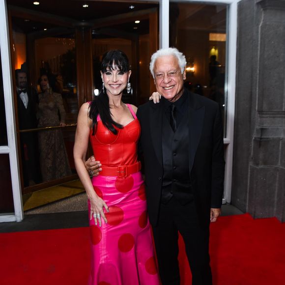 Alexandra Martins e Antônio Fagundes: Alexandra Martins veste um conjunto vibrante e estruturado, composto por um corset de couro vermelho com pespontos aparentes e uma saia longa em tom de rosa choque, decorada com grandes bolas (poás) em vermelho. O look é acentuado por um cinto largo que marca a cintura e brincos de cascata prateados. Ao seu lado, Antônio Fagundes opta por um visual monocromático preto, composto por terno, colete, camisa e gravata na mesma cor