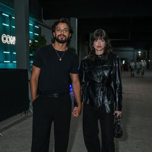 Rodrigo Simas e Agatha Moreira: Rodrigo veste um visual all black minimalista com camiseta básica de gola careca, calça de alfaiataria com cinto fino, sapatos sociais pretos, óculos de grau de armação fina e colar prateado; Agatha complementa a sintonia em tom escuro usando uma jaqueta de couro preta com textura craquelada, gola alta e amarração por cinto na cintura, combinada com calça ampla, bolsa preta com detalhes prateados e botas de bico quadrado