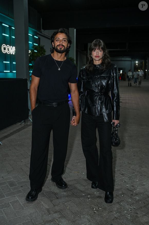 Rodrigo Simas e Agatha Moreira: Rodrigo veste um visual all black minimalista com camiseta básica de gola careca, calça de alfaiataria com cinto fino, sapatos sociais pretos, óculos de grau de armação fina e colar prateado; Agatha complementa a sintonia em tom escuro usando uma jaqueta de couro preta com textura craquelada, gola alta e amarração por cinto na cintura, combinada com calça ampla, bolsa preta com detalhes prateados e botas de bico quadrado