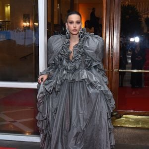 Livia Nunes roubou a cena com um dos looks mais dramáticos da noite, apostando em um vestido escultural em tafetá cinza chumbo. A peça é uma explosão de volumes, com maxi babados que contornam todo o decote em V profundo e se estendem pelos ombros, criando uma moldura teatral para o rosto. A saia volumosa, com camadas sobrepostas e textura levemente amassada, confere um ar de alta-costura vanguardista