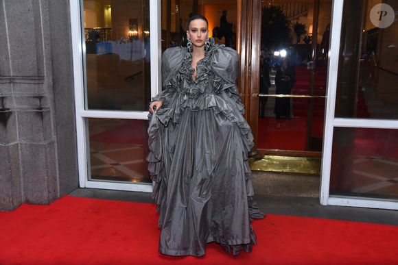 Livia Nunes roubou a cena com um dos looks mais dramáticos da noite, apostando em um vestido escultural em tafetá cinza chumbo. A peça é uma explosão de volumes, com maxi babados que contornam todo o decote em V profundo e se estendem pelos ombros, criando uma moldura teatral para o rosto. A saia volumosa, com camadas sobrepostas e textura levemente amassada, confere um ar de alta-costura vanguardista