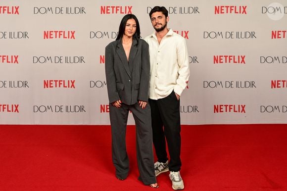 Yanna Lavigne aposta em um terninho oversized cinza chumbo com textura sutil. O visual é composto por um blazer de abotoamento simples e calças pantalona fluidas. Ela usa o blazer sem nada por baixo, criando um decote profundo, e completa com sandálias rasteiras pretas com tiras finas e pontas de metal dourado. Bruno Gissoni, usa um visual casual com camisa de botão off-white de linho amassado, calças cargo pretas e tênis esportivos cinzas com solado branco