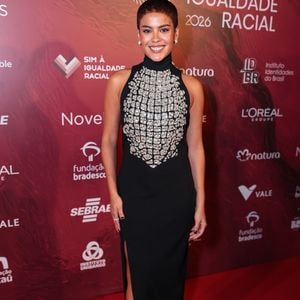 Bella Campos escolheu um visual minimalista e extremamente refinado, focado na silhueta e no detalhe frontal. A atriz veste um longo preto de gola alta e sem mangas, que traz como protagonista um bordado luxuoso de pedrarias prateadas em formato de escudo ou peitoral no centro do tronco. O corte seco do vestido, que conta com uma fenda lateral discreta, foi harmonizado com sapatos de bico fino e o cabelo curto bem rente à cabeça, valorizando seus traços naturais