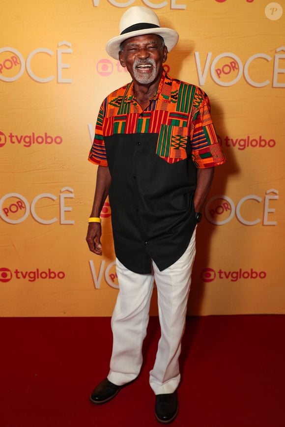 Antônio Pitanga esbanja elegância e brasilidade em uma camisa de mangas curtas com base preta e estampas étnicas multicoloridas no peito. O figurino é completado por calça de alfaiataria branca, sapatos sociais escuros e um clássico chapéu Panamá