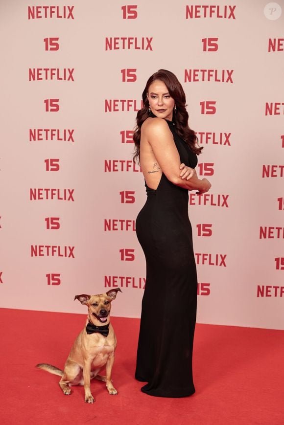 Paola Oliveira: traja um vestido longo preto, justo ao corpo e sem mangas, com gola alta estilizada e cava ampla que deixa parte das costas e da lateral do tórax à mostra; ela surge acompanhada de um cão com gravata borboleta preta; o caramelo fofo da Netflix roubou a cena ao lado da diva